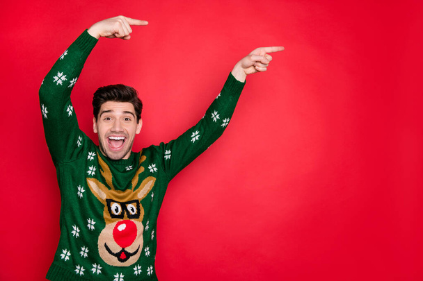 Photo of emotional handsome guy indicating fingers on x-mas sale banner wear trendy knitted green pullover with deer isolated red color background - 写真・画像