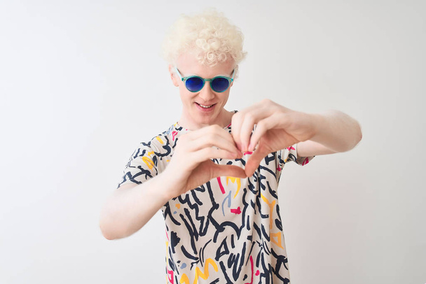Молодой белокурый альбинос в разноцветной футболке и солнцезащитных очках на изолированном красном фоне улыбается в любви, показывая символ и форму сердца руками. Романтическая концепция
. - Фото, изображение