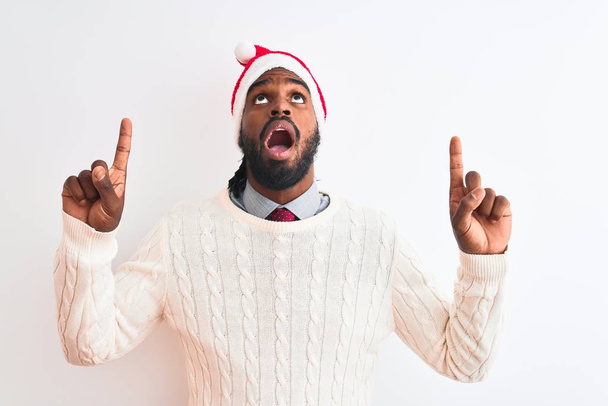 Jeune homme afro-américain portant le chapeau de Noël Santa sur fond blanc isolé étonné et surpris regardant vers le haut et pointant avec les doigts et les bras levés
. - Photo, image
