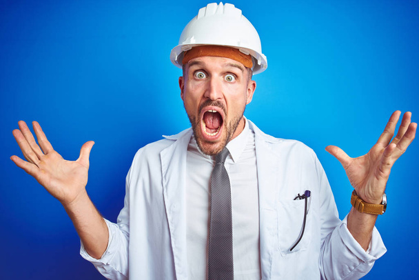 Young handsome engineer man wearing safety helmet over blue isolated background celebrating crazy and amazed for success with arms raised and open eyes screaming excited. Winner concept - Photo, Image