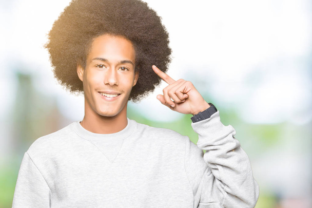 Молодой африканский американец с афроволосами в спортивной толстовке, улыбающийся, указывая пальцем на голову, отличная идея или мысль, хорошая память
 - Фото, изображение