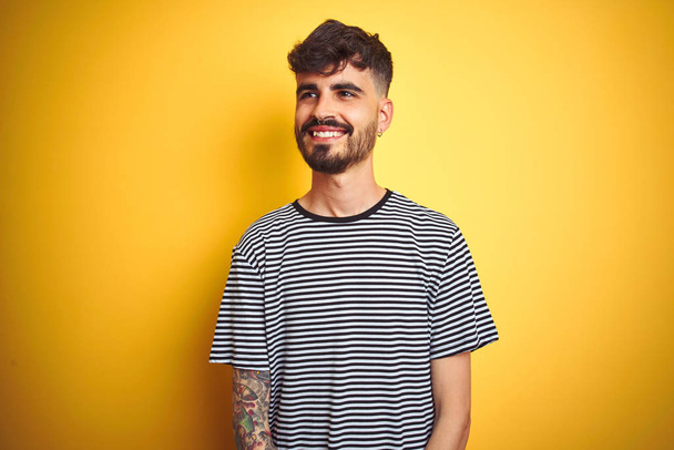 Young man with tattoo wearing striped t-shirt standing over isolated yellow background looking away to side with smile on face, natural expression. Laughing confident. - Photo, Image