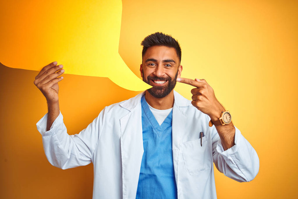 Young indian doctor man holding speech bubble standing over isolated yellow background very happy pointing with hand and finger - Photo, Image