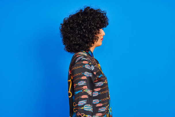 Young arab woman with curly hair wearing colorful shirt over isolated blue background looking to side, relax profile pose with natural face with confident smile. - Fotografie, Obrázek