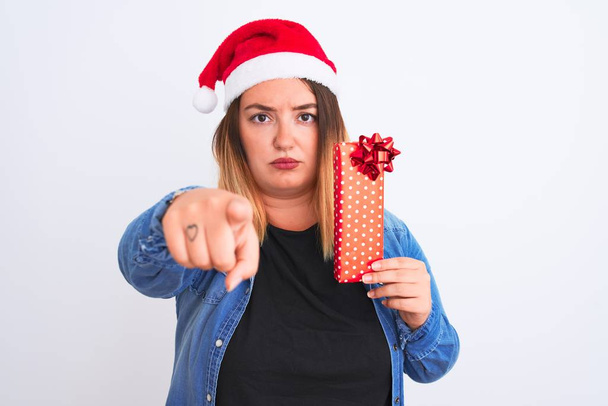 Piękna kobieta w kapeluszu Christmas Santa trzymając prezent na odizolowanym białym tle wskazując palcem na aparat i do Ciebie, znak ręczny, pozytywny i pewny gest z przodu - Zdjęcie, obraz