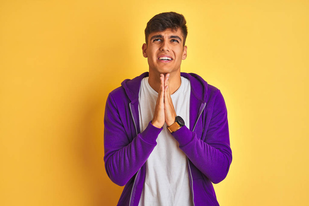Hombre joven indio con sudadera púrpura de pie sobre un fondo amarillo aislado mendigando y rezando con las manos juntas con expresión de esperanza en la cara muy emotiva y preocupada. Pedir perdón. Concepto de religión
. - Foto, imagen