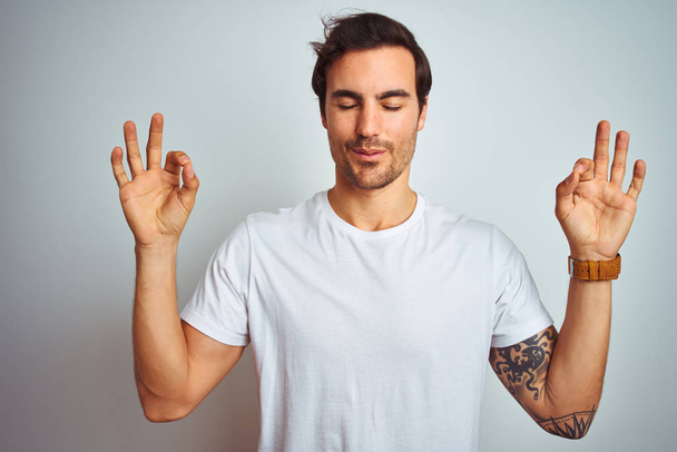 Young handsome man with tattoo wearing casual t-shirt over isolated white background relax and smiling with eyes closed doing meditation gesture with fingers. Yoga concept. - Foto, Bild