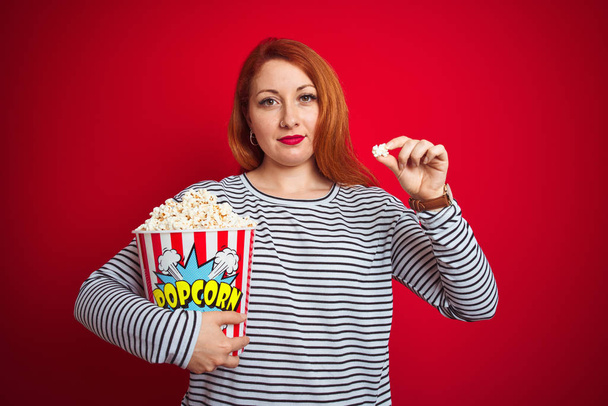 Mladý krásný ryšavý žena jíst popcorn přes červené izolované pozadí s jistým výrazem na inteligentní tvář myšlení vážné - Fotografie, Obrázek