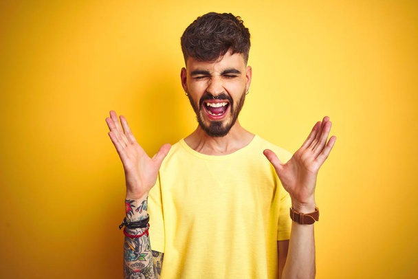 Молодой человек с татуировкой в футболке стоит на изолированном желтом фоне празднует безумный и безумный успех с поднятыми руками и закрытыми глазами, крича взволнованно. Концепция победителя
 - Фото, изображение