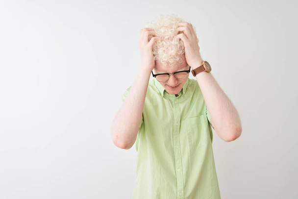 junger Albino-blonder Mann mit grünem Hemd und Brille über isoliertem weißem Hintergrund, der verzweifelt und gestresst unter Kopfschmerzen und Migräne leidet. Hände auf den Kopf. - Foto, Bild