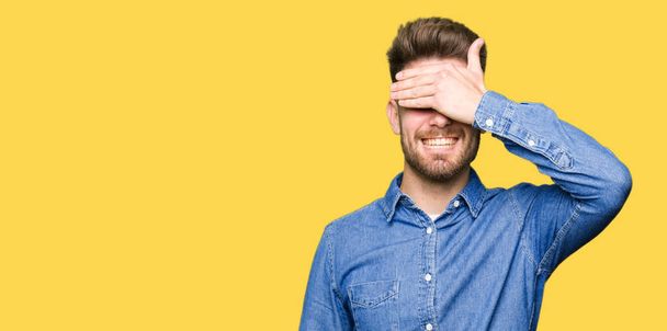 Young handsome blond man wearing casual denim shirt smiling and laughing with hand on face covering eyes for surprise. Blind concept. - 写真・画像