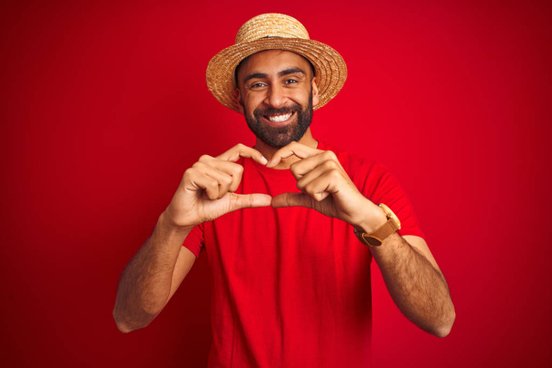 Joven hombre indio guapo usando camiseta y sombrero sobre fondo rojo aislado sonriendo en el amor mostrando el símbolo del corazón y la forma con las manos. Concepto romántico
. - Foto, Imagen