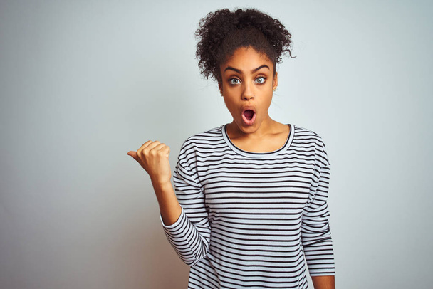Mujer afroamericana vistiendo camiseta de rayas navy de pie sobre fondo blanco aislado Sorprendido señalando con el dedo de la mano a un lado, boca abierta expresión asombrada
. - Foto, imagen