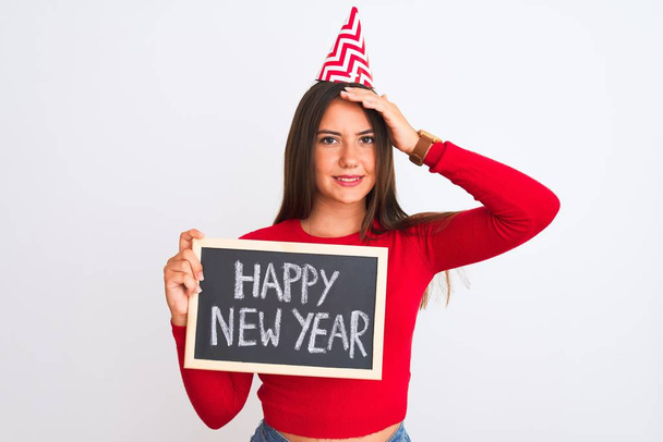 Beautiful girl wearing fanny party hat holding blackboard over isolated white background stressed with hand on head, shocked with shame and surprise face, angry and frustrated. Fear and upset for mistake. - Foto, Bild