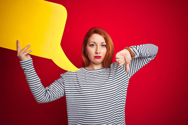 Mladá krásná zrzka žena drží žlutou řečovou bublinu nad červeným izolovaným pozadím s rozzlobeným obličejem, negativní znamení ukazující odpor s palci dolů, odmítavý koncept - Fotografie, Obrázek