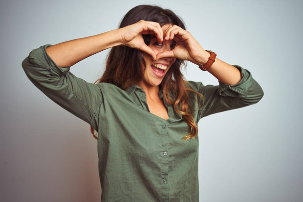 junge schöne Frau in grünem Hemd steht über grauem isoliertem Hintergrund und macht Herzform mit lächelnden Händen und Fingern, die durch Zeichen schauen - Foto, Bild