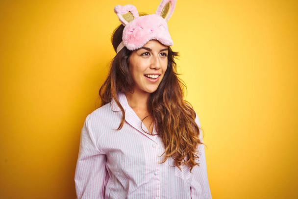 Young woman wearing pajama and sleep mask standing over yellow isolated background looking away to side with smile on face, natural expression. Laughing confident. - Photo, Image