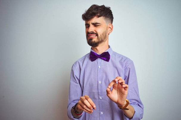 Молодий чоловік з татуюванням носить фіолетову сорочку та краватку на ізольованому білому тлі огидний вираз, незадоволений та боїться робити огидне обличчя, тому що реакція відрази. З піднятими руками. Концепція, що пропонує
. - Фото, зображення