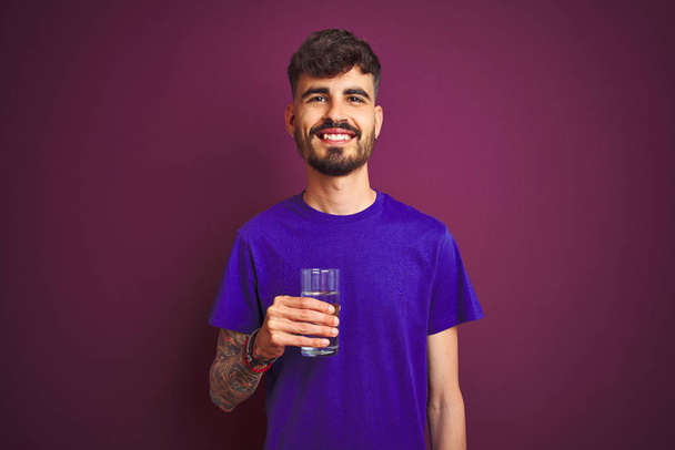 junger Mann mit Tätowierung, der ein Glas Wasser trinkt, das über einem isolierten lila Hintergrund steht, mit einem glücklichen Gesicht steht und mit einem selbstbewussten Lächeln lächelt, das Zähne zeigt - Foto, Bild