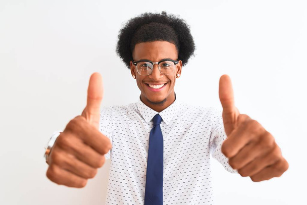 Joven hombre de negocios afroamericano con corbata y gafas sobre fondo blanco aislado que aprueba hacer un gesto positivo con la mano, pulgares arriba sonriendo y feliz por el éxito. Gesto del ganador
. - Foto, imagen