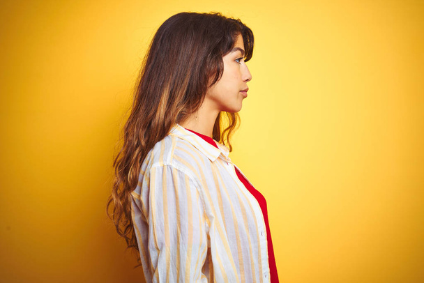 Jovem mulher bonita vestindo camiseta vermelha e listras camisa sobre fundo isolado amarelo olhando para o lado, relaxar pose perfil com rosto natural com sorriso confiante
. - Foto, Imagem