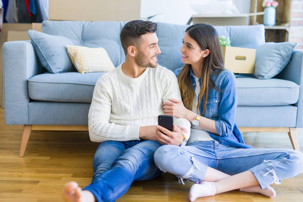 Jovem casal sentado no chão da nova casa usando smartphone e sorrindo feliz por se mudar para uma nova casa
 - Foto, Imagem