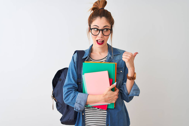 roodharige student vrouw dragen rugzak met boeken over geïsoleerde witte achtergrond wijzen en tonen met duim naar de zijkant met gelukkig gezicht glimlachen - Foto, afbeelding