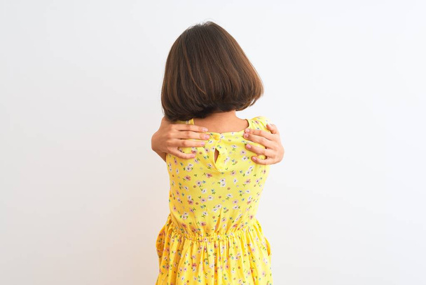 Jovem menina bonita usando vestido floral amarelo em pé sobre fundo branco isolado Abraçando-se feliz e positivo de trás para frente. Auto amor e auto cuidado
 - Foto, Imagem