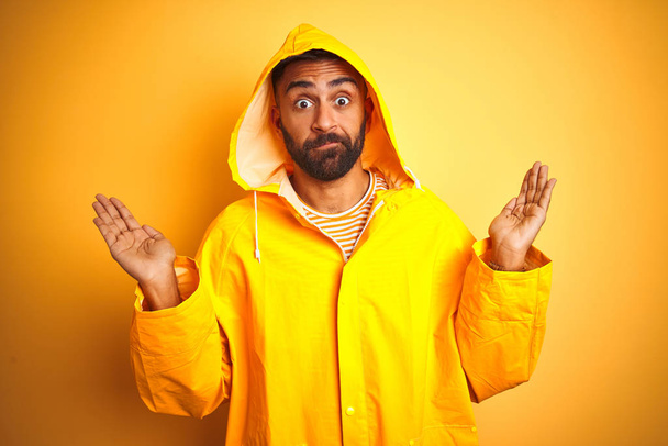 Jeune homme indien portant un imperméable avec capuche debout sur fond jaune isolé expression inutile et confus avec les bras et les mains levées. Concept de doute
. - Photo, image