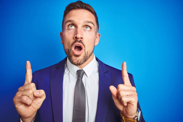 Close up picture of young handsome business man over blue isolated background amazed and surprised looking up and pointing with fingers and raised arms. - Photo, Image