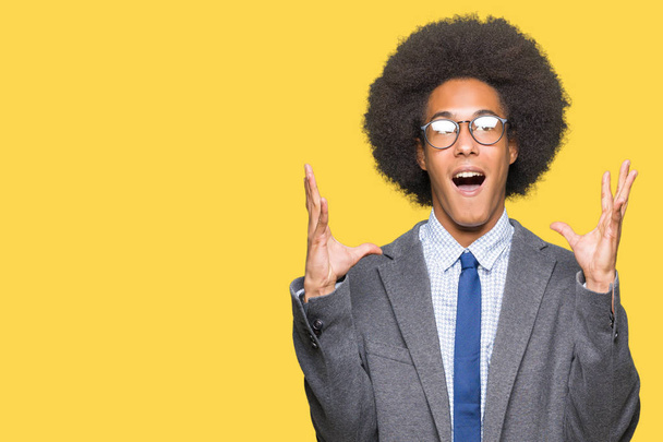 Jovem empresário afro-americano com cabelo afro vestindo óculos celebrando loucos e maravilhado pelo sucesso com braços levantados e olhos abertos gritando animados. Conceito de vencedor
 - Foto, Imagem