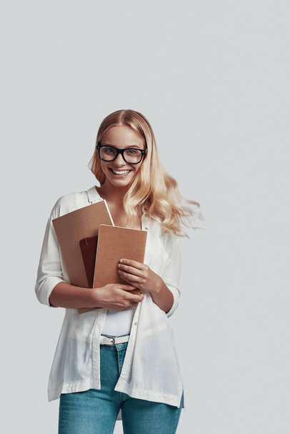 Vonzó fiatal nő szemüveg könyv és mosolyog, miközben áll szemben szürke háttér - Fotó, kép