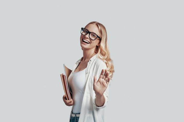 Ελκυστική νεαρή γυναίκα με γυαλιά που κουβαλάει βιβλία και χαμογελά ενώ στέκεται στο γκρίζο φόντο - Φωτογραφία, εικόνα