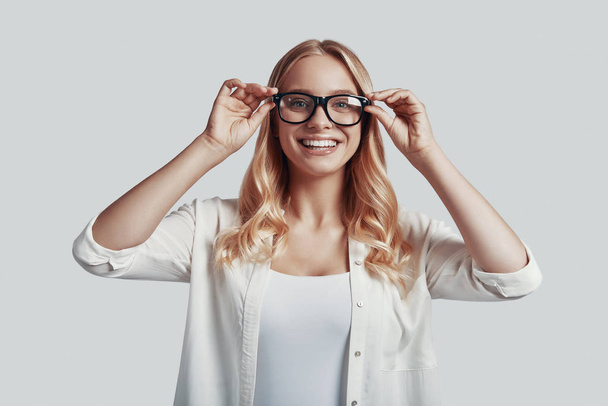 Ελκυστική νεαρή γυναίκα προσαρμόζει τα γυαλιά και χαμογελά ενώ στέκεται πάνω σε γκρι φόντο - Φωτογραφία, εικόνα