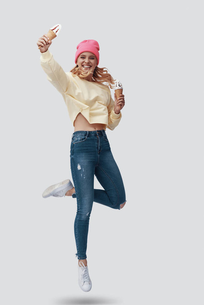 Full length of attractive young woman eating ice cream and smiling while hovering against grey background - Foto, Imagem
