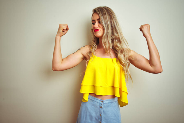 Mujer hermosa joven con camiseta amarilla de pie sobre fondo blanco aislado que muestra los músculos de los brazos sonriendo orgulloso. Concepto Fitness
. - Foto, imagen