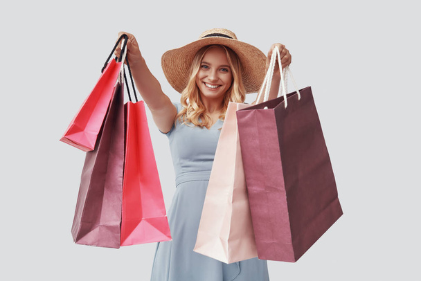Atraktivní mladá žena dívá na kameru s úsměvem a nesoucí nákupní tašky, zatímco stojí na šedém pozadí  - Fotografie, Obrázek