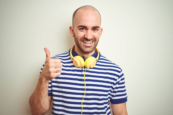 Mladý muž, který poslouchá hudbu ve žlutých sluchátkách na izolovaném pozadí, dělá šťastnou ruku s rukou. Povolování výrazu s pohledem na fotoaparát s úspěšným zobrazením. - Fotografie, Obrázek