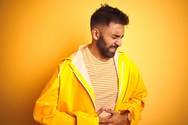 Young indian man wearing raincoat standing over isolated yellow background with hand on stomach because indigestion, painful illness feeling unwell. Ache concept. - Photo, Image