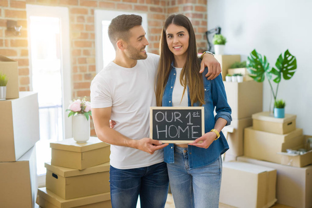 Όμορφο νεαρό ζευγάρι που κινείται σε ένα νέο σπίτι, χαμογελαστός πολύ χαρούμενος κρατώντας πίνακα με το πρώτο μας κείμενο στο σπίτι - Φωτογραφία, εικόνα