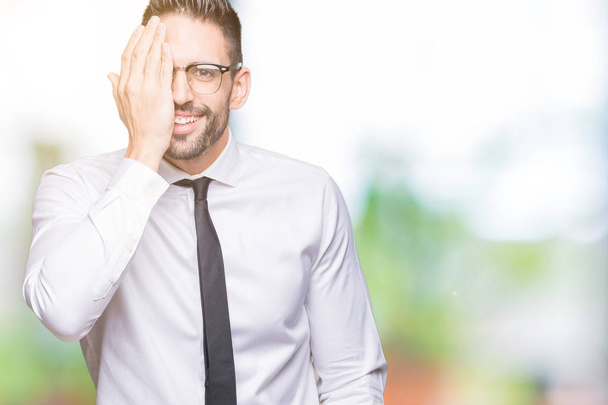 Young handsome business man wearing glasses over isolated background covering one eye with hand with confident smile on face and surprise emotion. - 写真・画像
