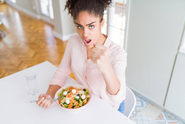 Jonge Afro-Amerikaanse vrouw eten gezonde Pasta Salade geïrriteerd en gefrustreerd schreeuwen met woede, gek en schreeuwen met verhoogde hand, woede concept - Foto, afbeelding