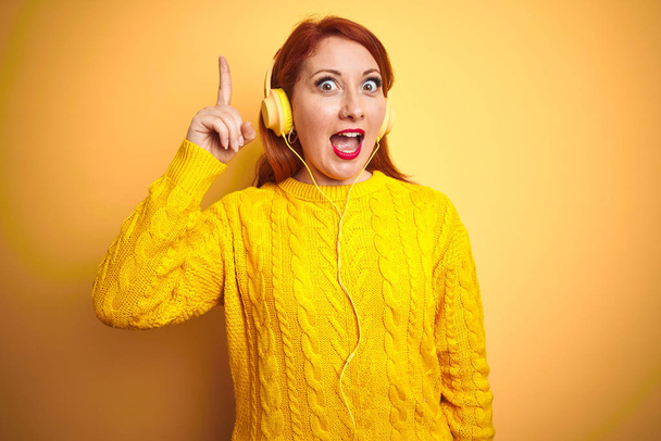 Νεαρή κοκκινομάλλα γυναίκα που ακούει μουσική χρησιμοποιώντας ακουστικά πάνω από κίτρινο απομονωμένο φόντο δείχνοντας το δάχτυλο επάνω με επιτυχημένη ιδέα. Έξοδος και ευτυχία. Νούμερο ένα.. - Φωτογραφία, εικόνα