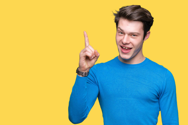 Молодой красивый мужчина в синем свитере на изолированном фоне указывает пальцем вверх с успешной идеей. Взволнован и счастлив. Номер один
. - Фото, изображение