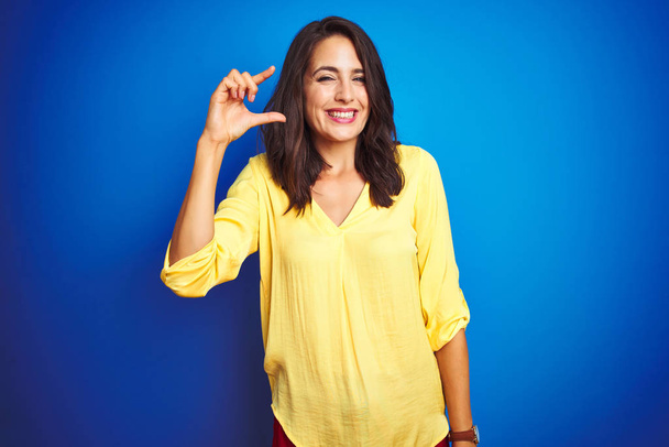 Jonge mooie vrouw draagt gele t-shirt staan over blauw geïsoleerde achtergrond glimlachend en zelfverzekerd gebaar met de hand doen kleine formaat teken met vingers kijken en de camera. Maatconcept. - Foto, afbeelding