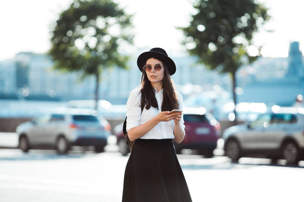 young woman in a hat and a white shirt walks in the city and uses a smartphone. - Foto, Bild