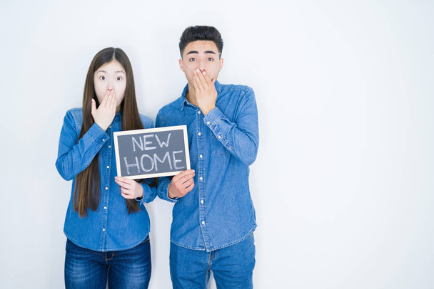 Beautiful young asian couple over white background holding blackboard with new home text cover mouth with hand shocked with shame for mistake, expression of fear, scared in silence, secret concept - Photo, Image