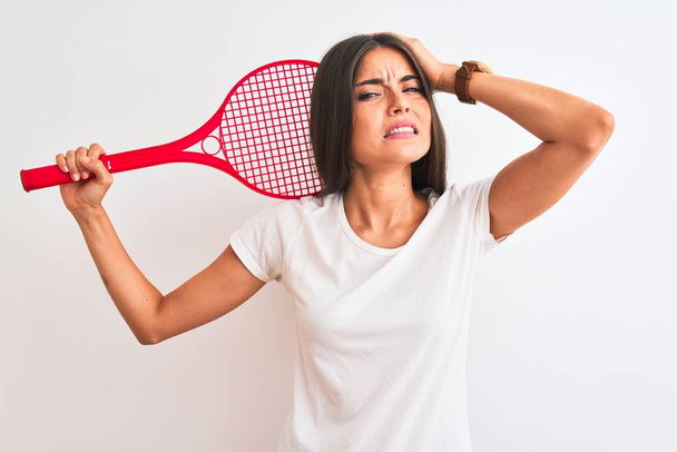 Красива спортсменка грає в теніс, використовуючи ракетку, що стоїть над ізольованим білим тлом, напруженим рукою на голові, шокована соромом і несподіваним обличчям, сердитим і розчарованим. Страх і засмучення за помилку
. - Фото, зображення