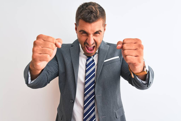 Young handsome business man wearing suit and tie over isolated background angry and mad raising fists frustrated and furious while shouting with anger. Rage and aggressive concept. - Foto, Imagem