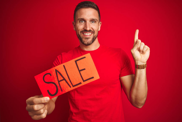 Young man holding sale advertising poster board over red isolated background very happy pointing with hand and finger to the side - Fotografie, Obrázek